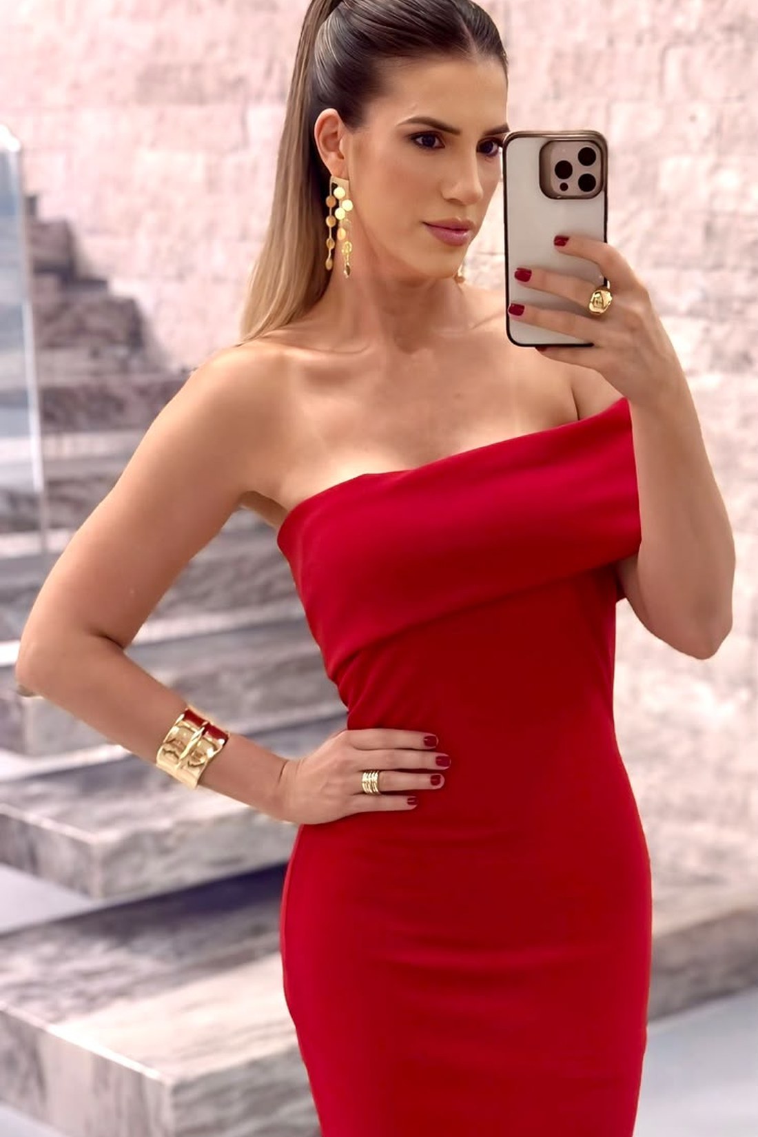 Fafá de vestido vermelho ombro único, com brincos e bracelete dourados, numa escadaria de pedra clara